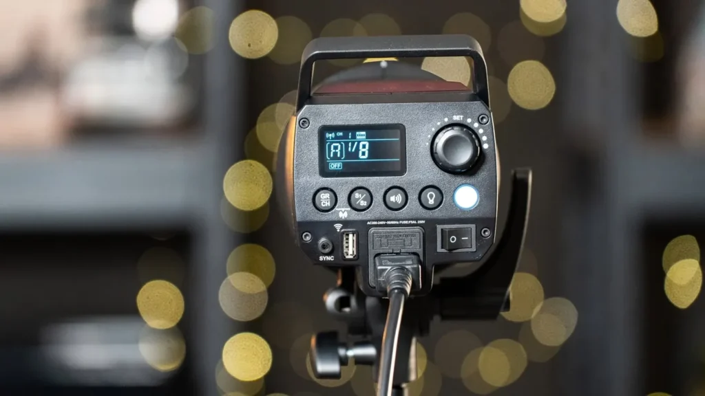 Задня панель моноблока Godox MS300-V на стійці з підключеним мережевим кабелем, екран показує потужність 1/8, канал 1, на розмитому фоні вогники боке Задня панель моноблока Godox MS300-V на стійці з підключеним мережевим кабелем, екран показує потужність 1/8, канал 1, на розмитому фоні вогники боке